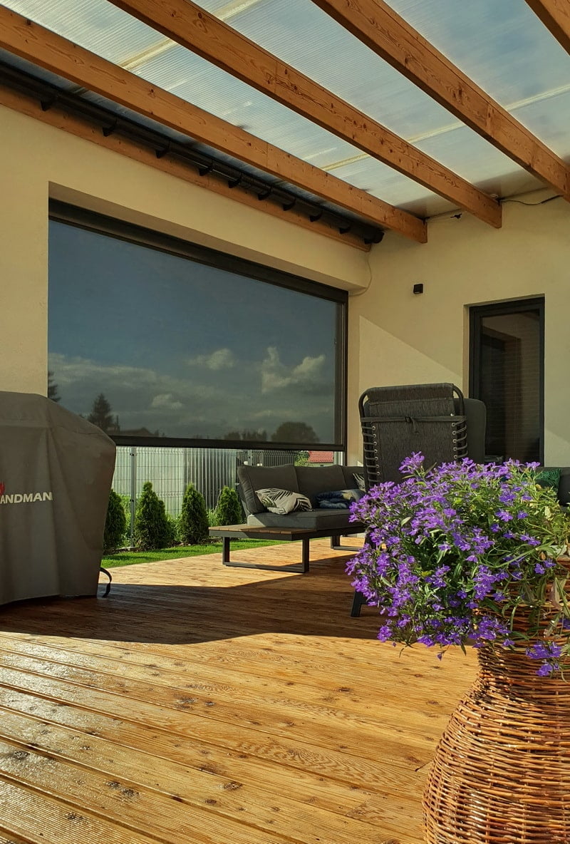 Wooden deck patio with purple flowers in a woven basket, outdoor seating, and a covered grill beside a large glass door under a translucent roof.