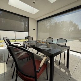 Outdoor dining table with six black chairs on a covered patio, set with plates and bread, shaded by retractable screens.