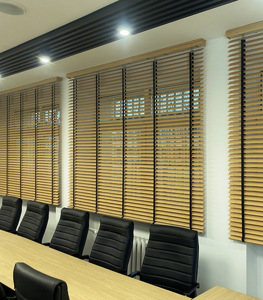 Conference room with a long wooden table and black cushioned chairs, windows covered by horizontal wooden blinds.