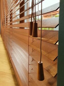Close-up of brown Venetian blinds with pull cords and weighted tassels hanging inside a sunlit window area