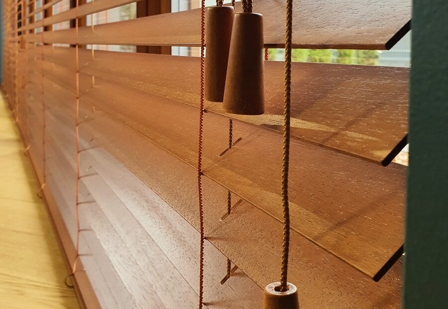 Close-up of brown Venetian blinds with pull cords and weighted tassels hanging inside a sunlit window area
